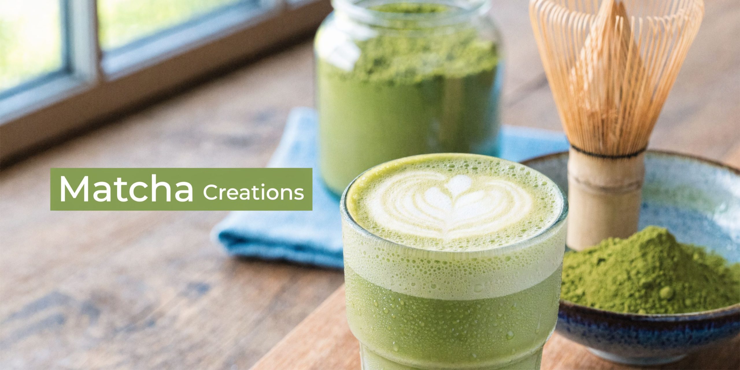 A fresh matcha latte with latte art next to a bamboo whisk and matcha powder