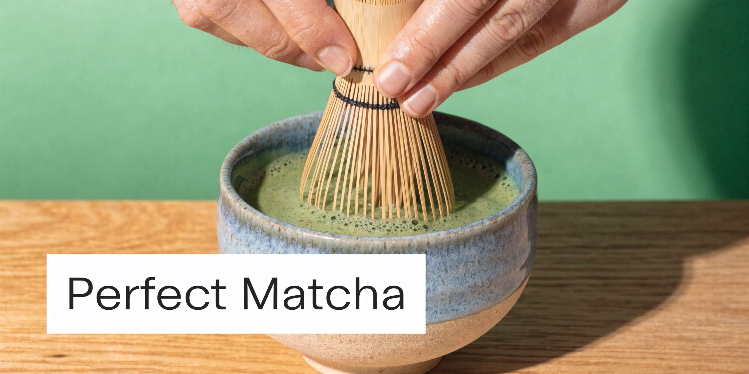 A pair of hands whisking green matcha tea in a ceramic bowl with a bamboo whisk.