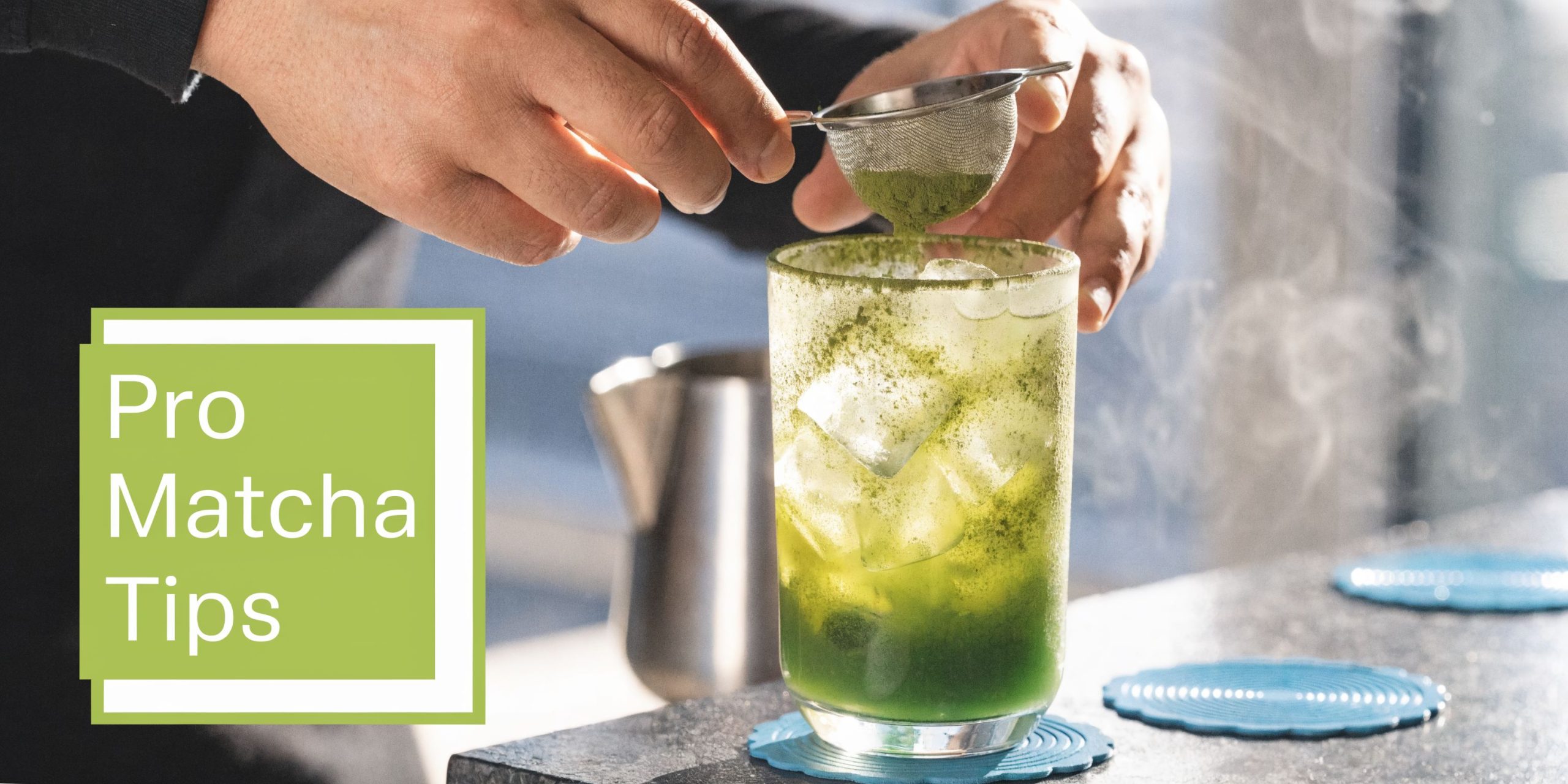 A person uses a small metal sieve to dust matcha powder over a glass of iced tea.