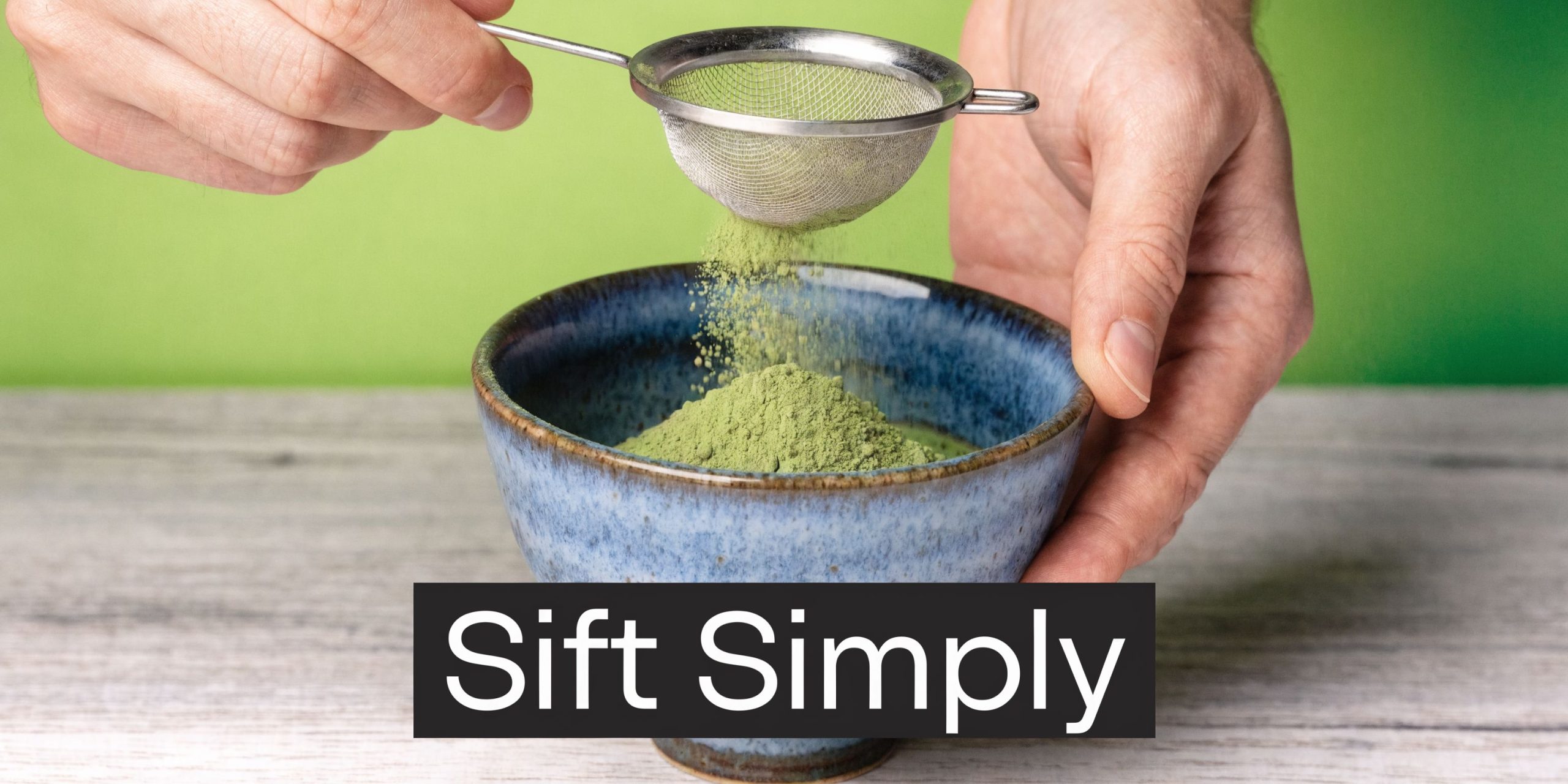 A person sifting vibrant green matcha powder into a blue ceramic bowl on a wooden surface.