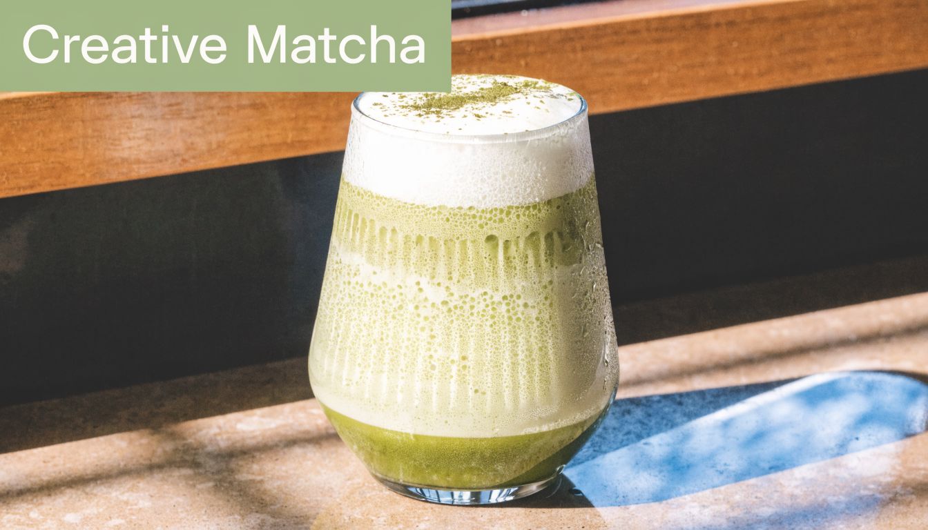 A glass of iced matcha latte with creamy foam topping sitting on a sunny marble surface.