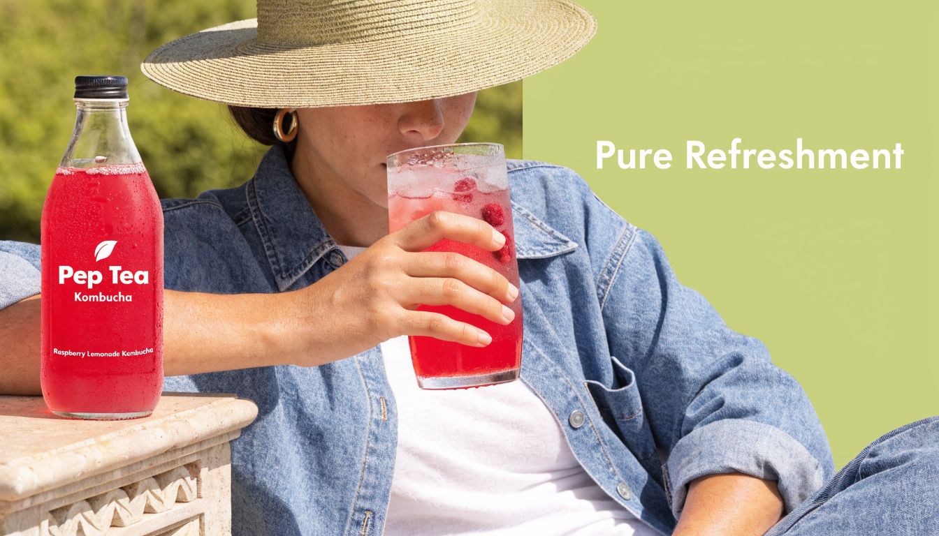 A woman wearing a straw hat drinks raspberry lemonade kombucha from a glass while relaxing outdoors.