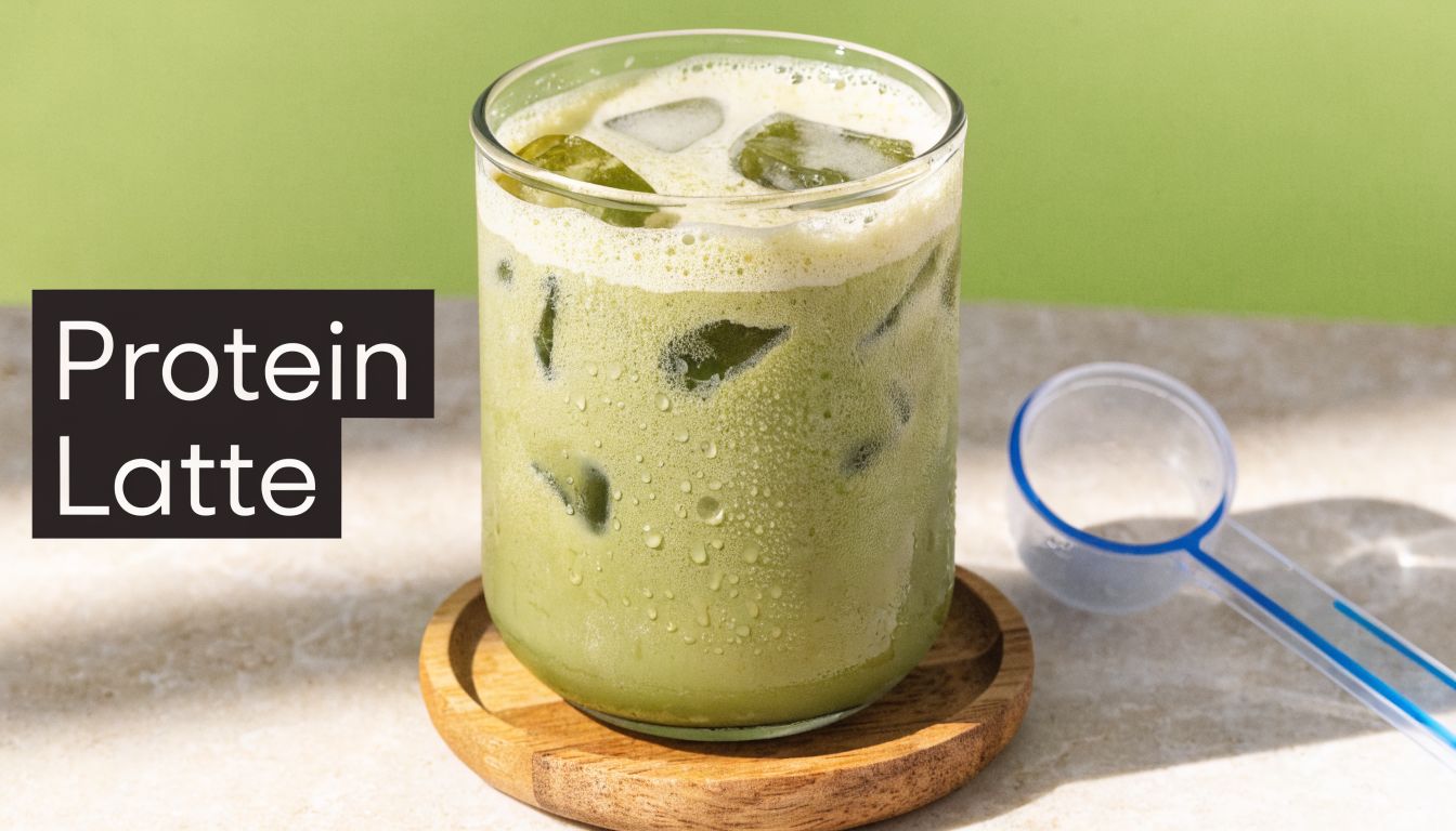 A refreshing iced green matcha protein latte in a clear glass on a wooden coaster.