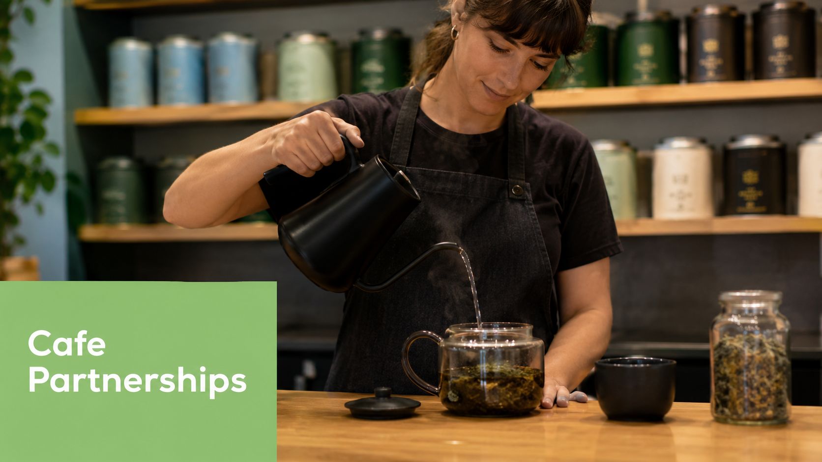A professional barista pouring hot water into a glass teapot filled with loose leaf tea in a cafe.