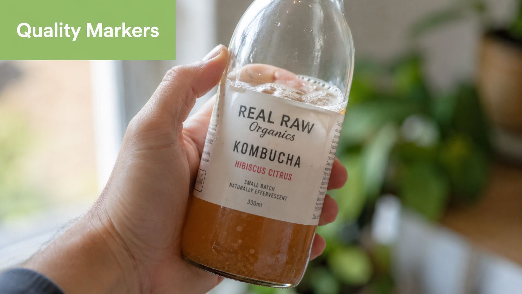 A hand holding a glass bottle of Real Raw Organics Hibiscus Citrus kombucha against a blurred background.