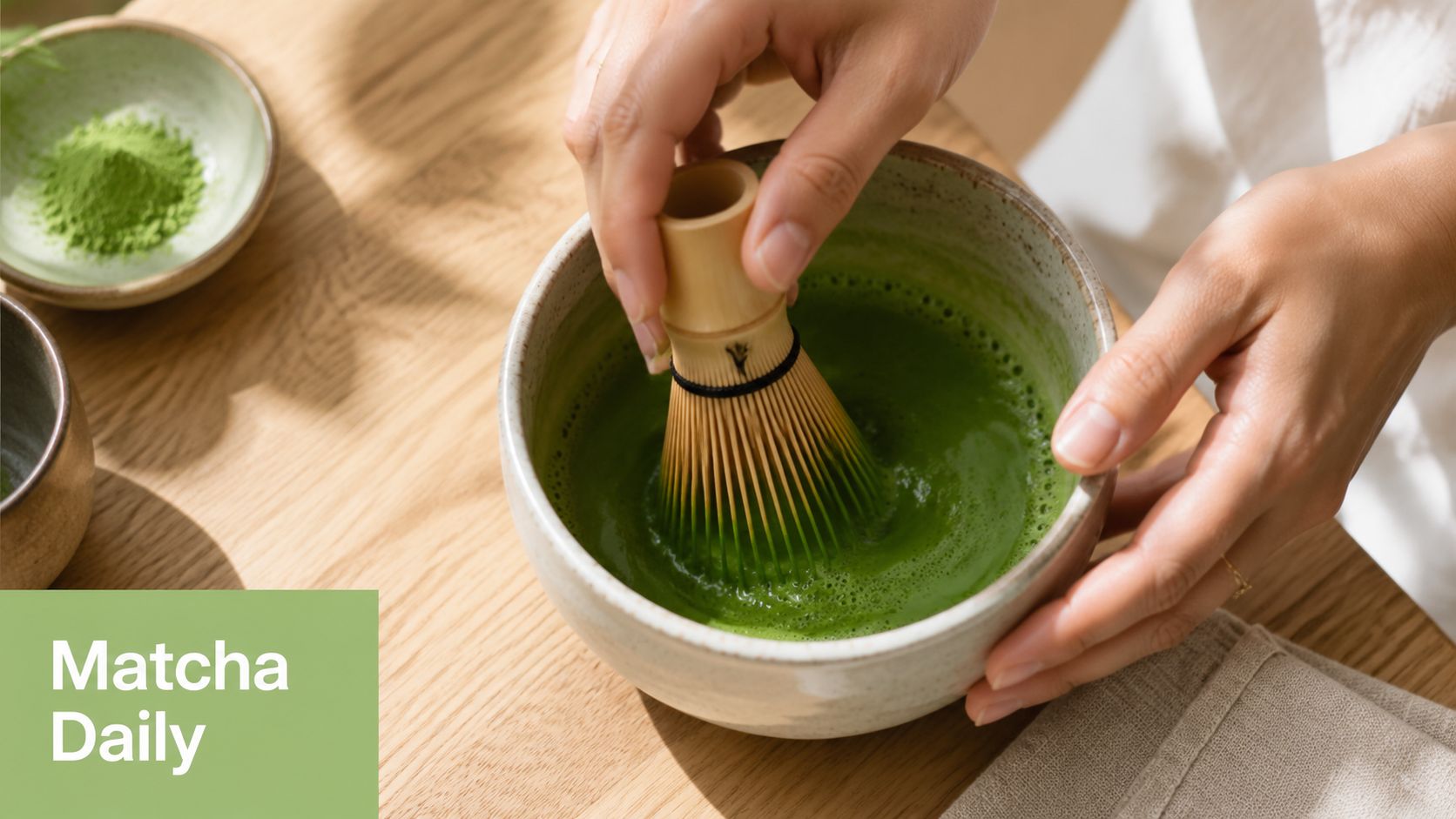 A person preparing matcha green tea with a bamboo whisk in a ceramic bowl on a table.